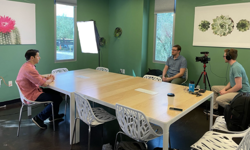 Two people interviewing one person on opposite sides of a large table with camera equipment.